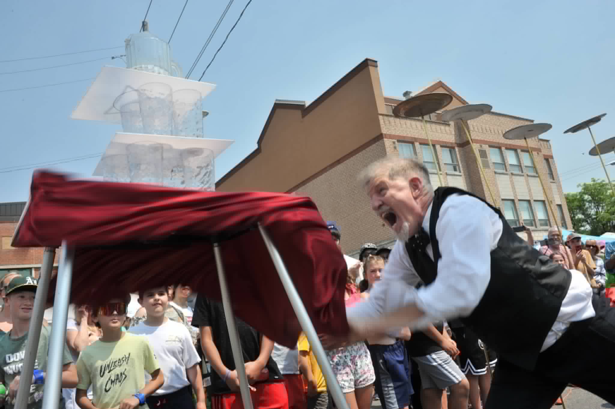 High-speed tablecloth yank under a tower of glasses at a street festival
